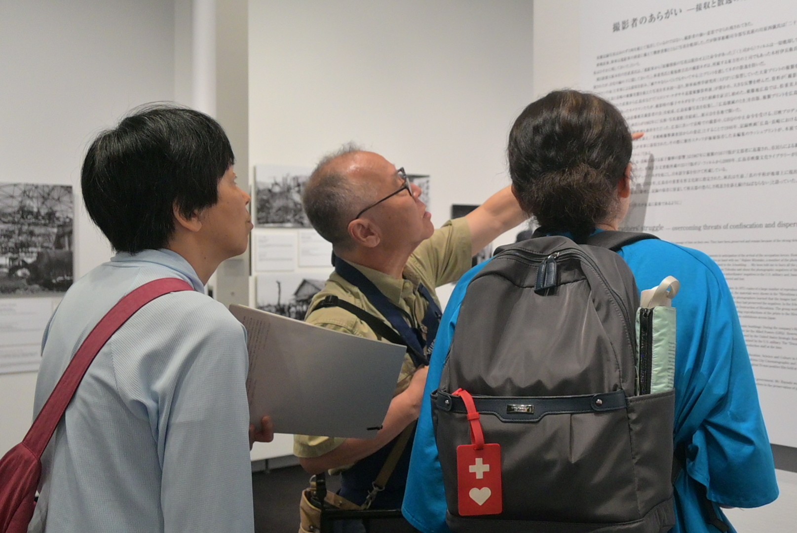 美術館の展示室内で、壁面に掲示された大きな解説パネルを3人の来場者が間近で読み込んでいる様子。中央の人物が解説文の一節を指さし、隣の2人がそれを熱心に見つめている。一人はリュックにヘルプマークをつけている。