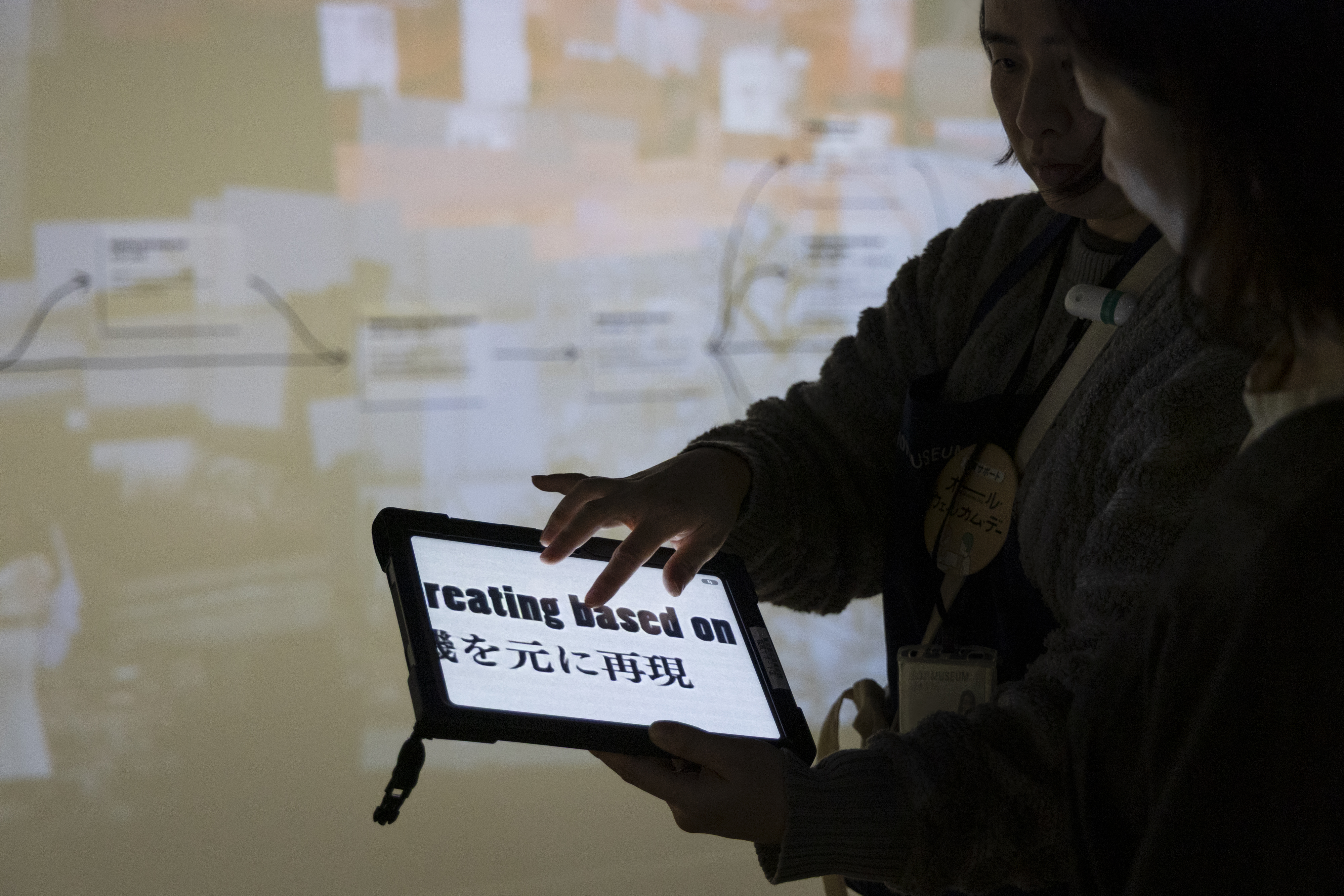 薄暗い展示空間で、スタッフがタブレット端末を操作している手元のアップ。画面には文字が大きく高コントラストで表示されている。