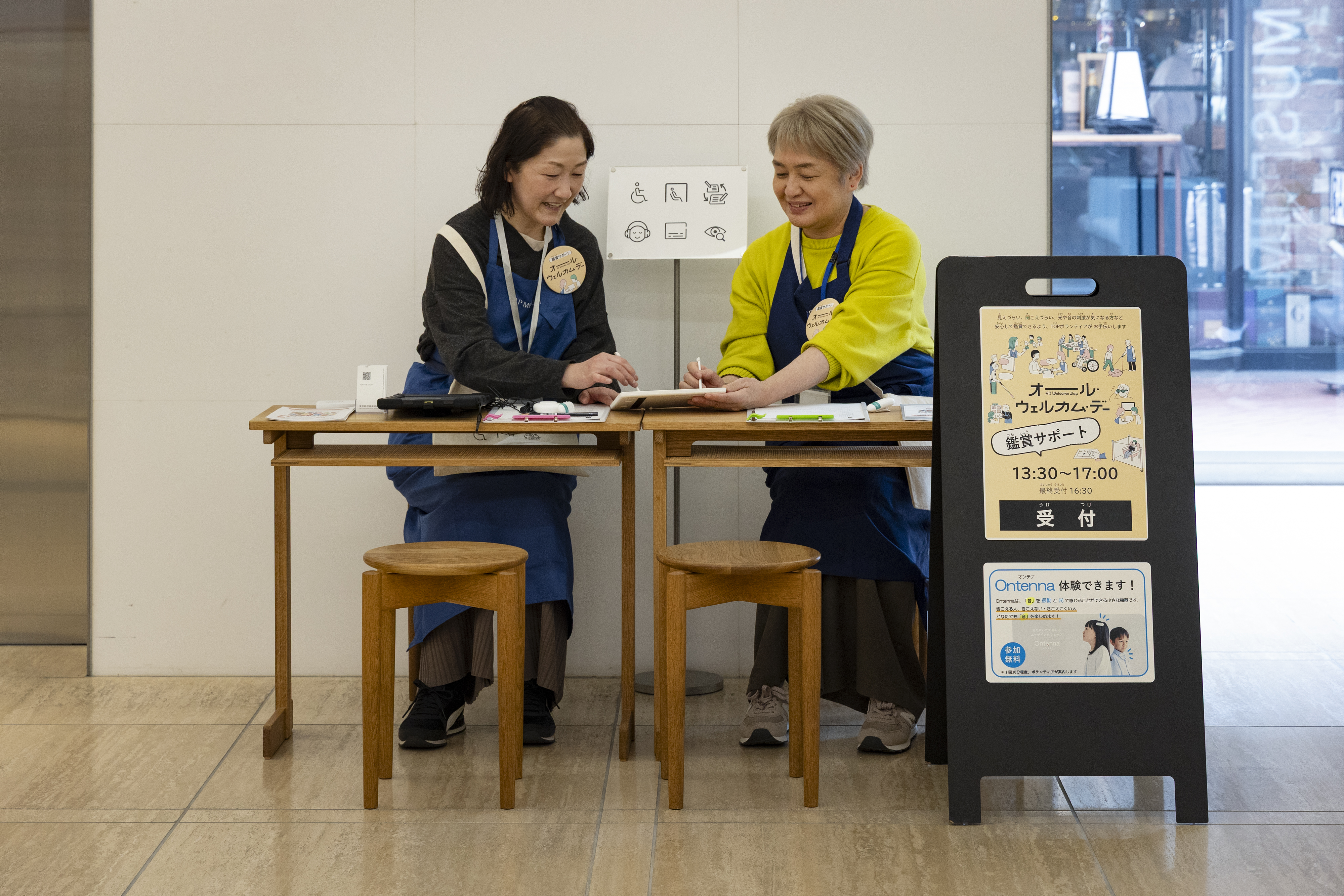 美術館の受付デスクに座る、お揃いの青いエプロンをつけた2人の女性スタッフ。デスクの横には「オール・ウェルカム・デー 受付」と書かれた看板があり、背後には車椅子や補聴器などのアクセシビリティに関するピクトグラムも表示されている。