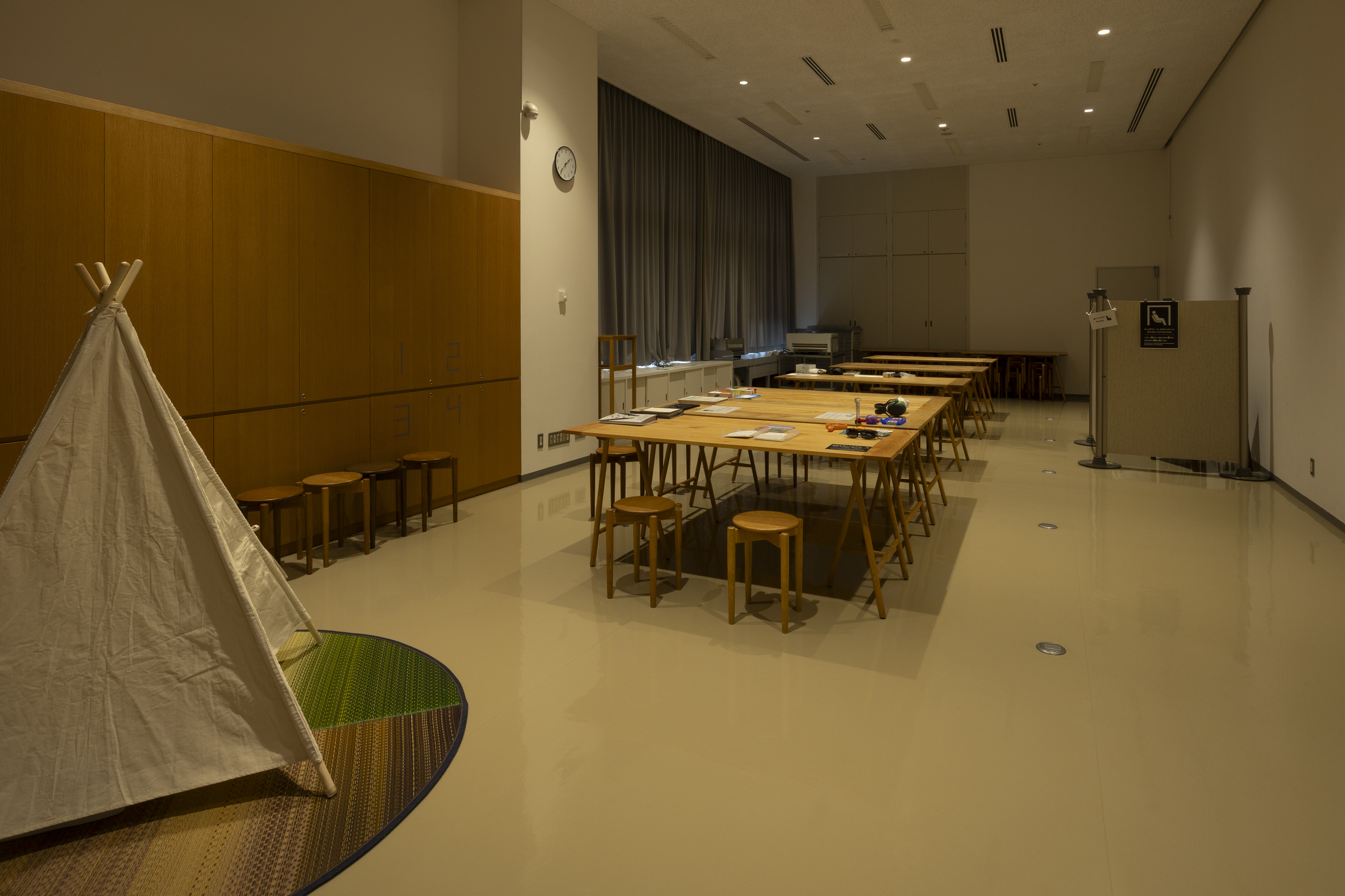 美術館の中の休憩のための部屋。中央には長い木製のテーブルと椅子が並び、手前には子供が遊べるような小さな白いティピーテントと円形のカラフルなラグが置かれている。奥にはパーテーションで仕切ったスペースもある。